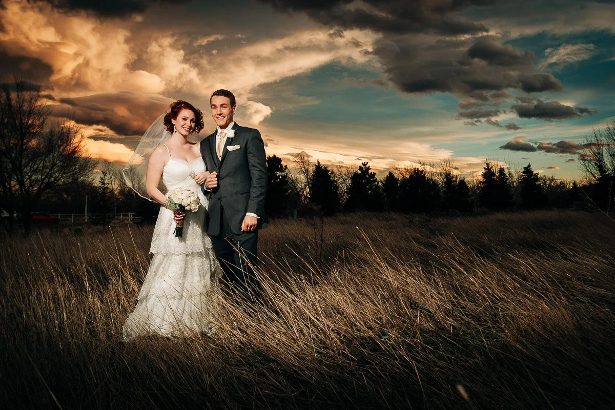 Bride and groom stand smiling in a field under a dramatic sunset sky. The bride holds a bouquet, and the scene conveys joy and romance.
