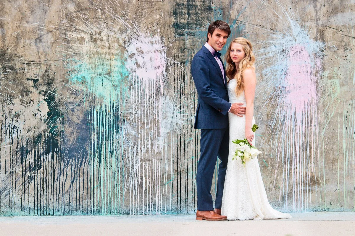 A bride in a white gown and groom in a blue suit stand embracing before an abstract, colorful wall. The scene conveys love and celebration.