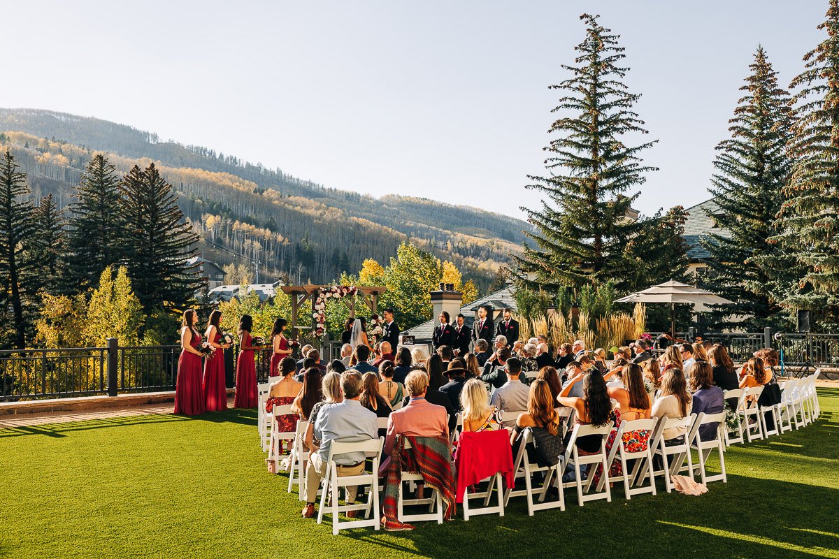 Outdoor Beaver Creek wedding ceremony on a sunny day features guests seated on white chairs facing the altar. Bridesmaids in burgundy dresses stand nearby, with mountains in the background.