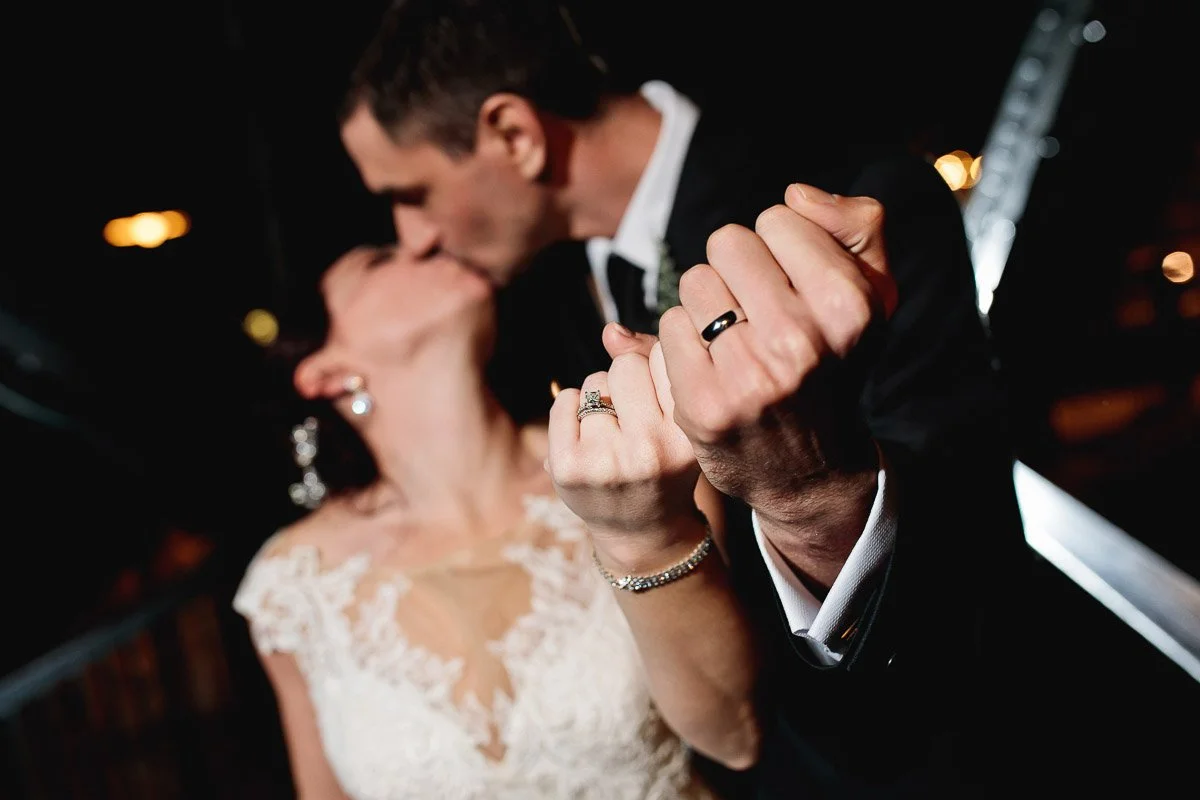 A bride and groom clasp pinkies and kiss. The focus is on their hands showing wedding rings, with the couple wearing elegant attire.