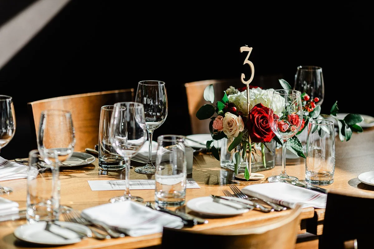 Elegant table setting with wood table, empty wine glasses, cutlery, white napkins, and a floral centerpiece with red and white roses on a dark background.