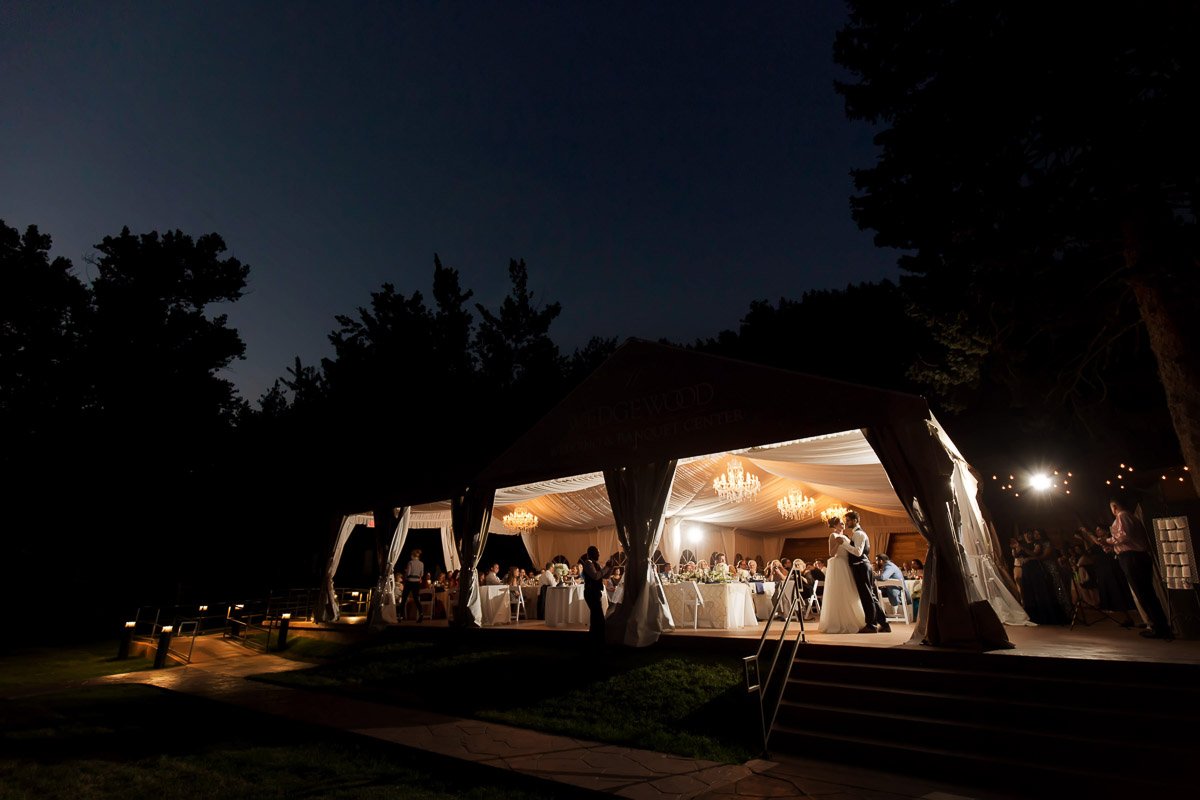 A glowing outdoor tent at dusk hosts a wedding reception. Inside, guests are seated at tables under warm chandeliers. A couple shares a dance, exuding joy.