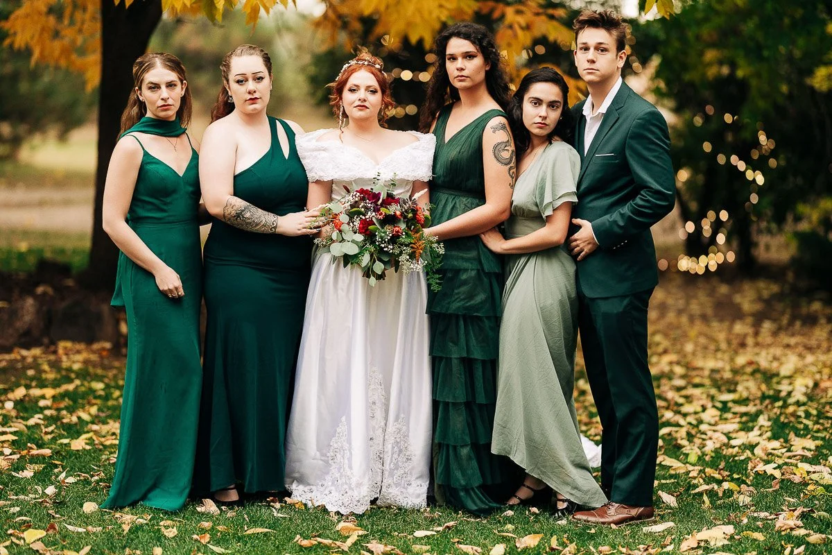 A group of six people, one in a white wedding dress holding a bouquet, stand solemnly on a leafy lawn. Others are in various green dresses and a suit.
