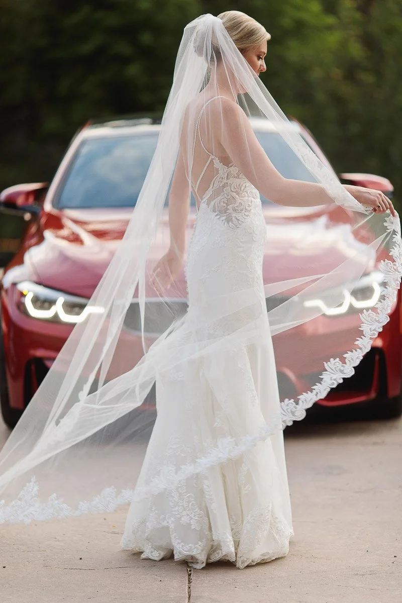 A bride in a white lace wedding dress and long veil stands gracefully in front of a red car, exuding elegance and joy against a natural backdrop.