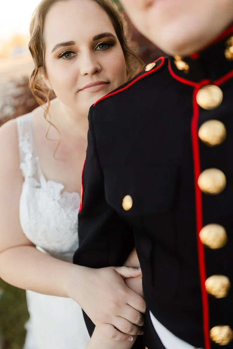 A bride in a white lace dress holds the arm of a person in a military uniform with gold buttons and red trim, conveying love and pride.