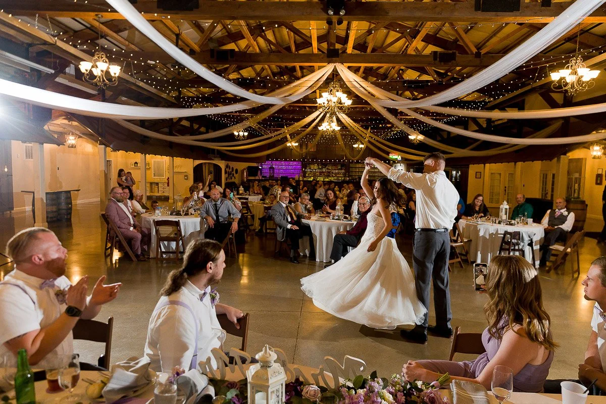 A couple dances joyfully in the center of a warmly lit reception hall, surrounded by seated guests. Draped ceiling and chandeliers add elegance.