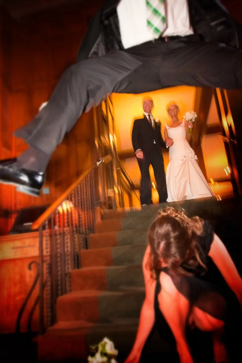 A playful wedding scene with a couple smiling on stairs. A man in a suit playfully leaps in the foreground as a woman picks up flowers below.