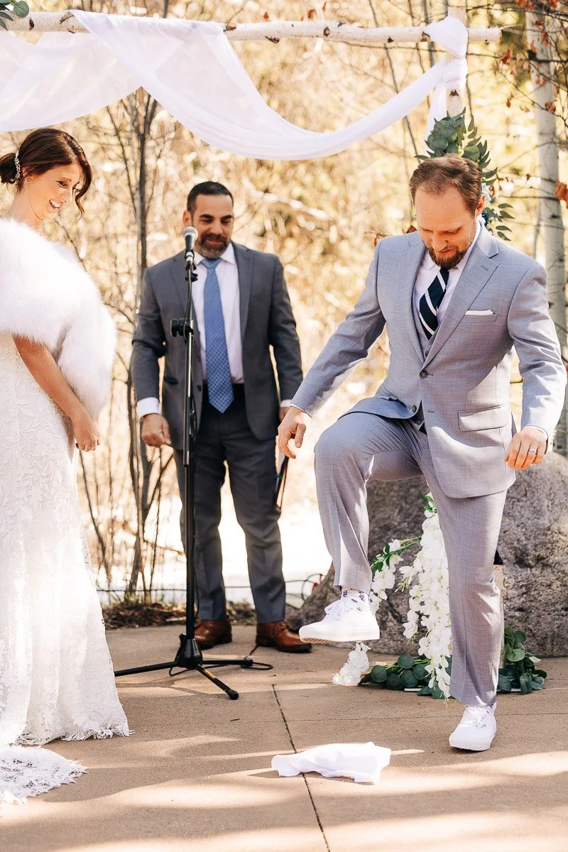 A groom in a light gray suit steps on a glass wrapped in cloth under a wedding arch. The bride, in a white gown and shawl, smiles at him. A man in a suit stands behind them.