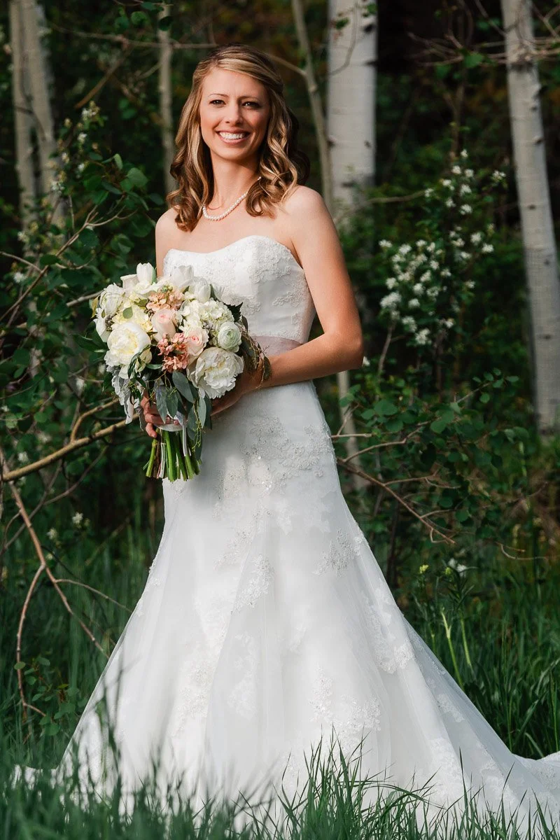 Bride in white lace gown holding a bouquet of white and pink flowers, standing in a lush forest with green foliage, smiling joyfully.
