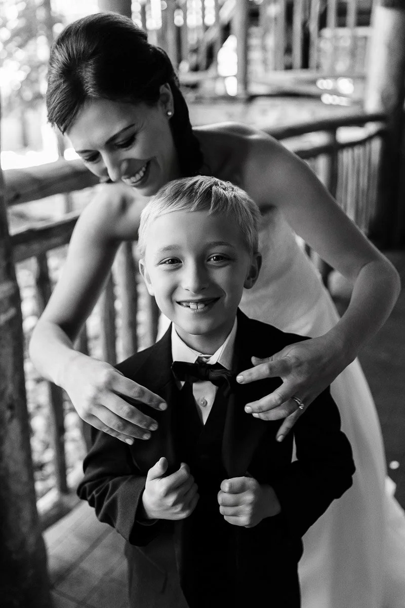 A smiling woman in a wedding dress hugs a young boy in a suit from behind on a wooden porch. The scene conveys joy and warmth.