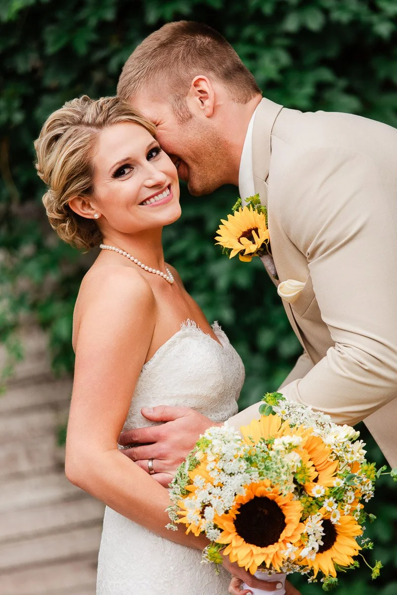 Bride in a strapless lace gown smiles while groom in beige whispers, holding sunflower bouquet. Green foliage backdrop adds romantic charm.