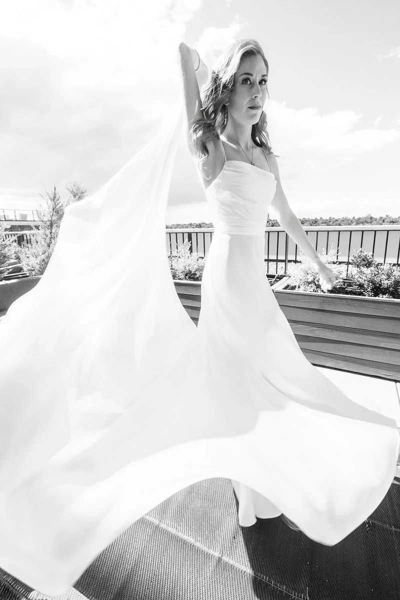 Woman in a flowing white dress twirls gracefully on a sunlit balcony, with a blurred background of sky and trees. The scene conveys elegance and freedom.