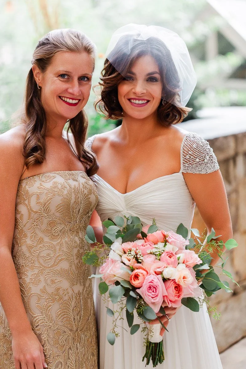Two smiling women in elegant dresses, one in a white bridal gown with a bouquet of pink and white roses, the other in a gold strapless dress.