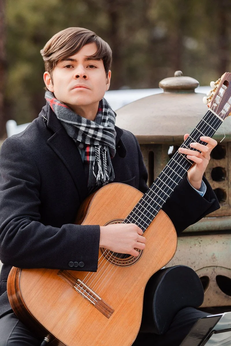 A young man sits outdoors holding a classical guitar. He wears a dark coat and plaid scarf, exuding a serious and focused expression. Forest background.