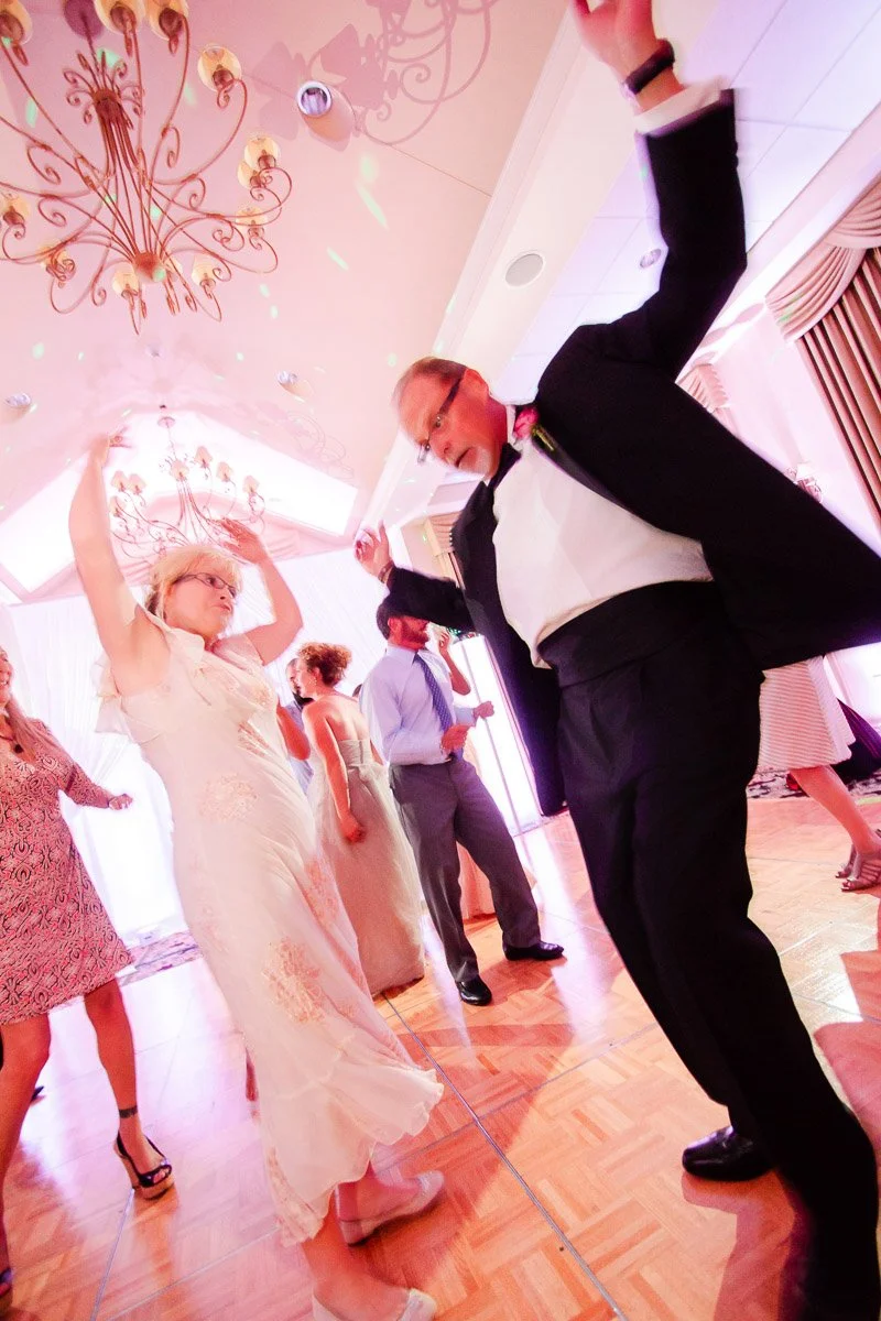 Guests dancing joyfully at a wedding reception. A woman in a white dress and a man in a tuxedo dance under ornate chandeliers, surrounded by others.