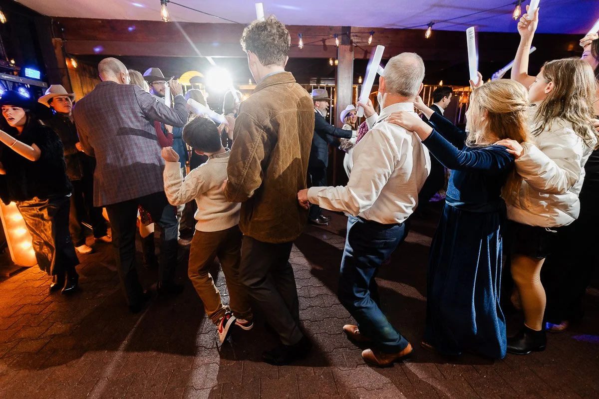 A lively group of people of various ages joyfully dance in a conga line at a party, under warm string lights. Some hold glowing sticks, enhancing the festive atmosphere.