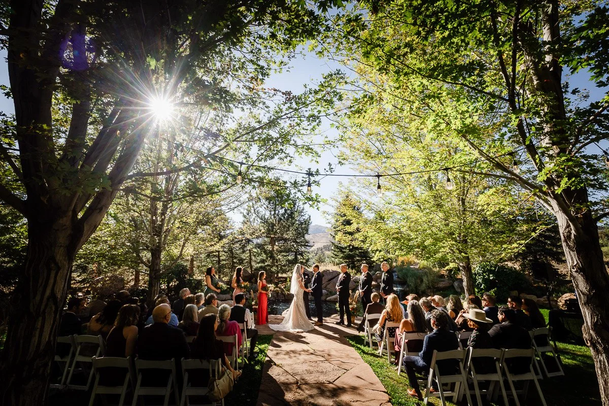A sunlit outdoor wedding ceremony under tall trees with guests seated on either side. The couple stands at the altar with bridesmaids and groomsmen nearby during a Greenbriar Inn wedding in Boulder, Colorado