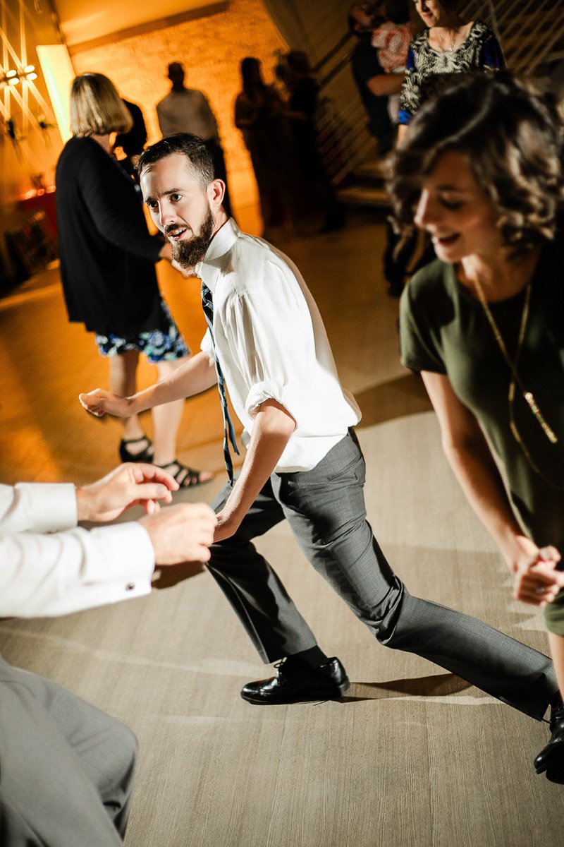 People dance energetically in a warmly lit room, conveying joy and excitement. A bearded man in a tie strikes a playful pose, smiling.