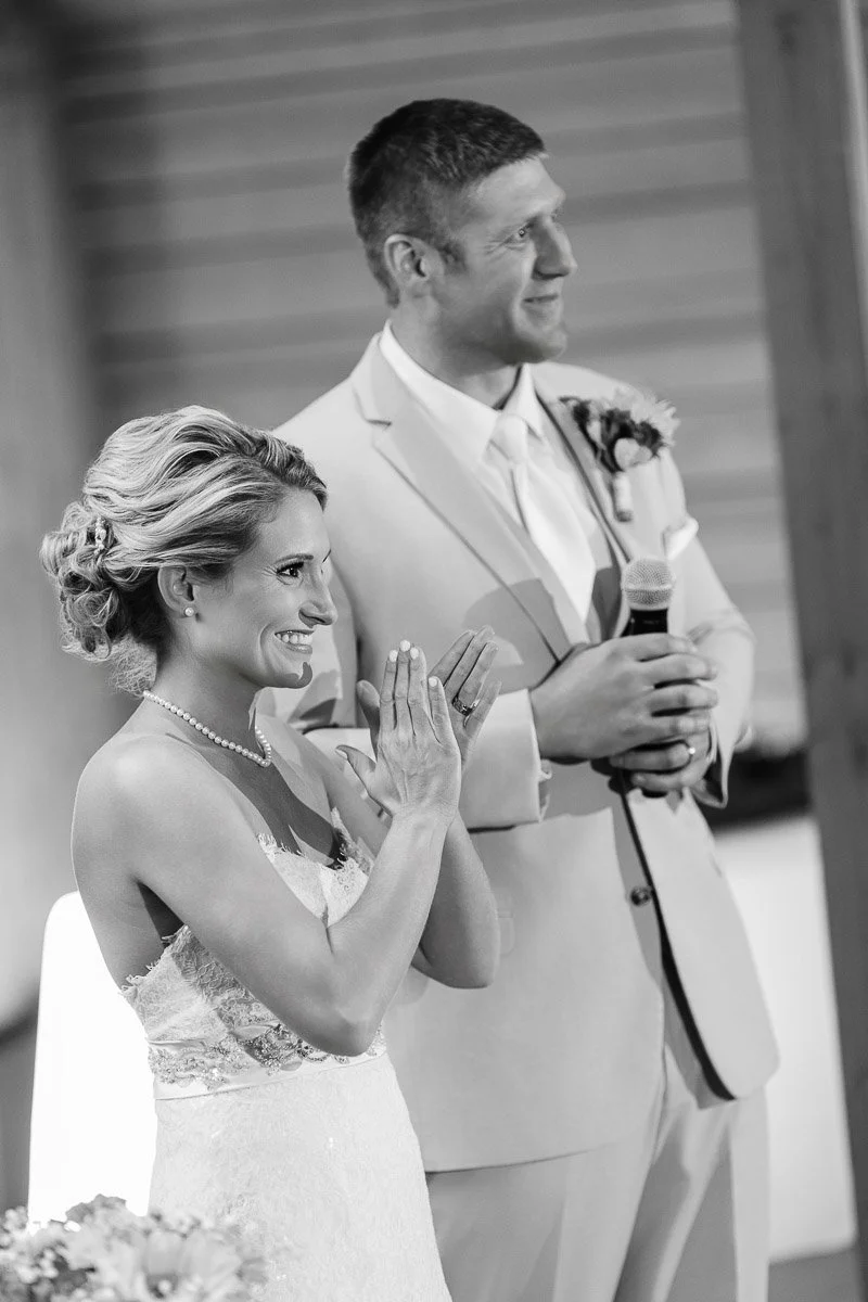 A joyful couple at their wedding; the woman, in a lace gown, clasps her hands in excitement, while the man in a suit smiles, holding a microphone.