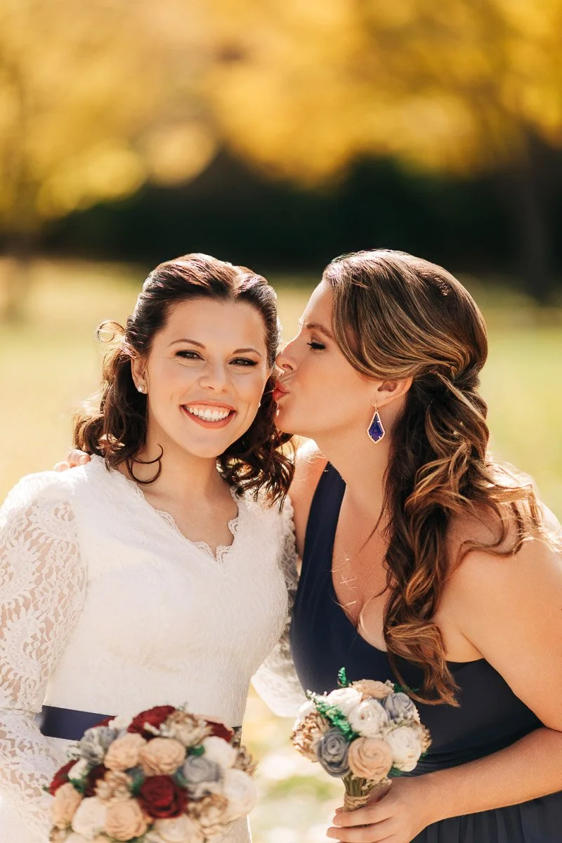 Two women smiling in a sunny, outdoor setting. The one on the left in a white lace dress holds a bouquet. Her companion in navy leans in for a kiss. Warm, joyful atmosphere.