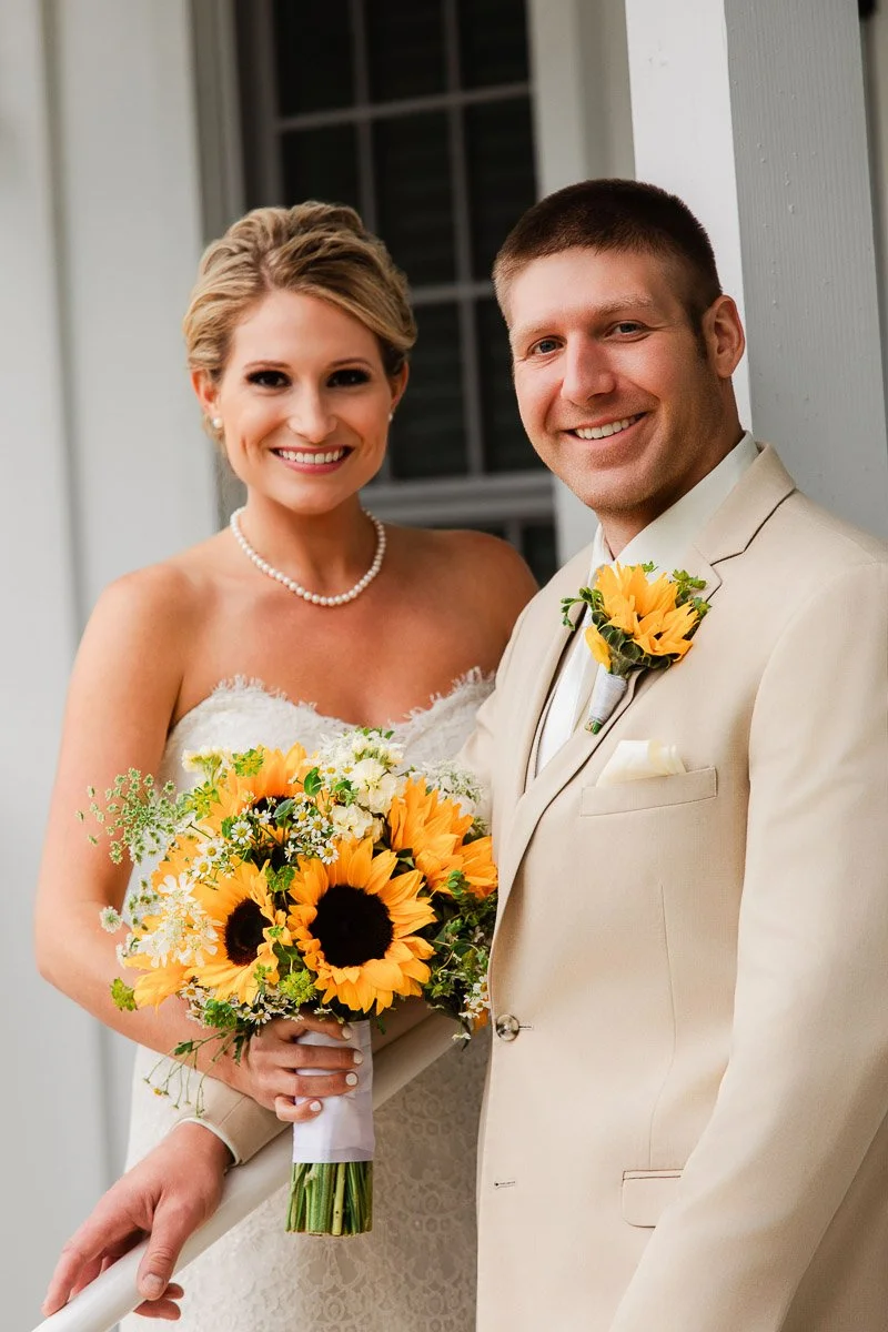Bride in a lace strapless gown holding a sunflower bouquet, standing beside a groom in a beige suit with a sunflower boutonniere, both smiling warmly.