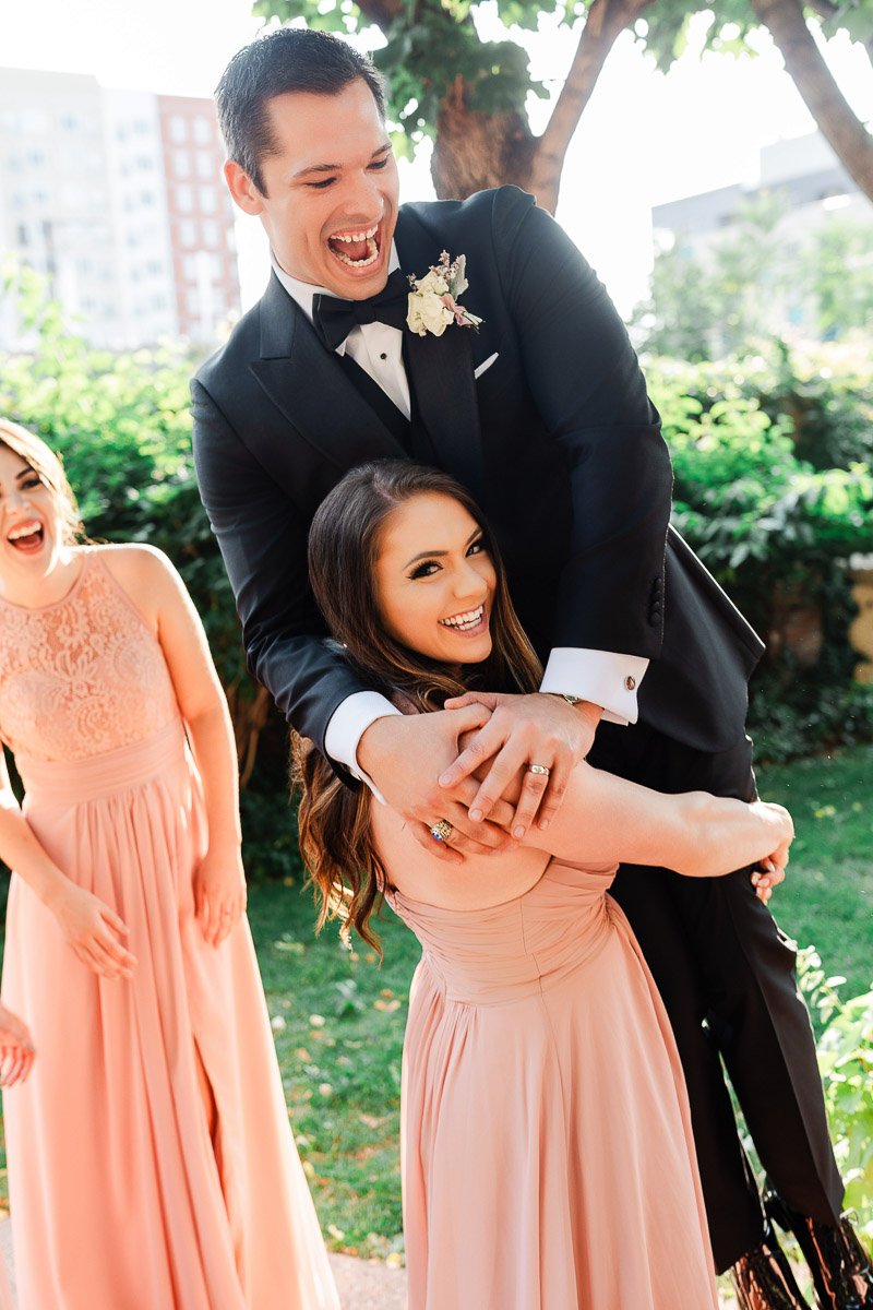 A joyful scene at an outdoor wedding; a woman in a pink dress playfully lifts a man in a black tuxedo. Another woman in pink laughs nearby.