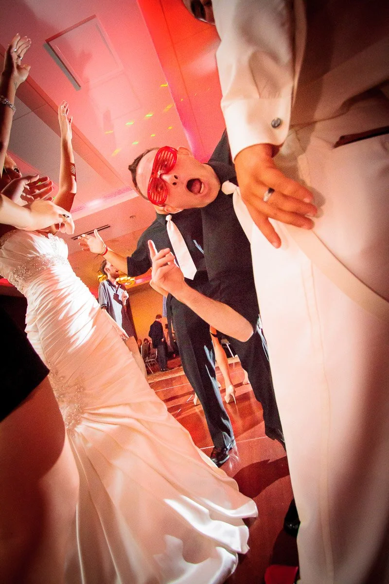 A lively wedding scene with people dancing. A man in red glasses and a black outfit makes a funny face, surrounded by others in formal attire. Joyous atmosphere.