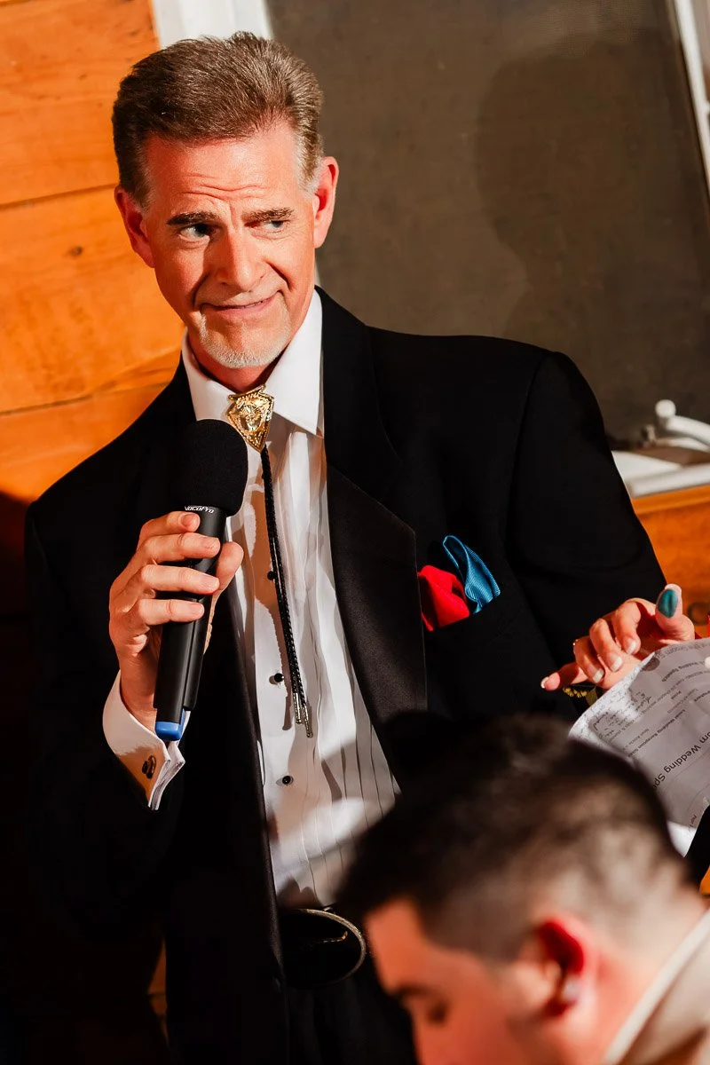 A confident man in a tuxedo, holding a microphone, gestures while speaking. He wears a decorative bolo tie and pocket square, exuding a formal and engaging atmosphere.