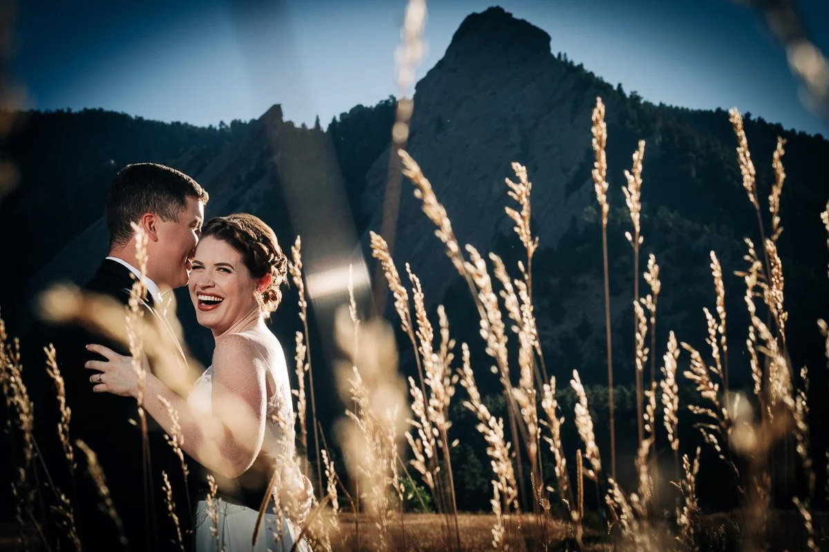 A joyful bride and groom stand in a field of tall golden grasses with The Flatirons. The bride laughs brightly, conveying happiness and celebration captured by Boulder Wedding Photographer tomKphoto