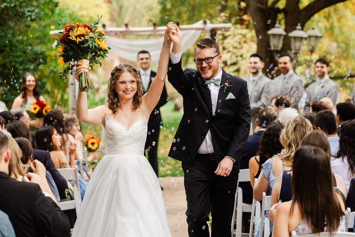 A joyful bride and groom hold hands, walking down an outdoor aisle surrounded by guests. The bride holds sunflowers; bubbles float in the air.