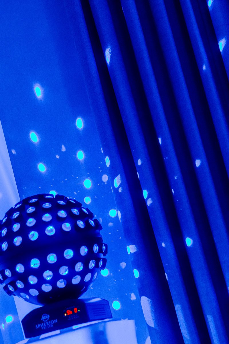 A dark disco ball casts vibrant blue dots on a curtain and wall, creating a lively, festive atmosphere. The scene exudes an energetic, party vibe.