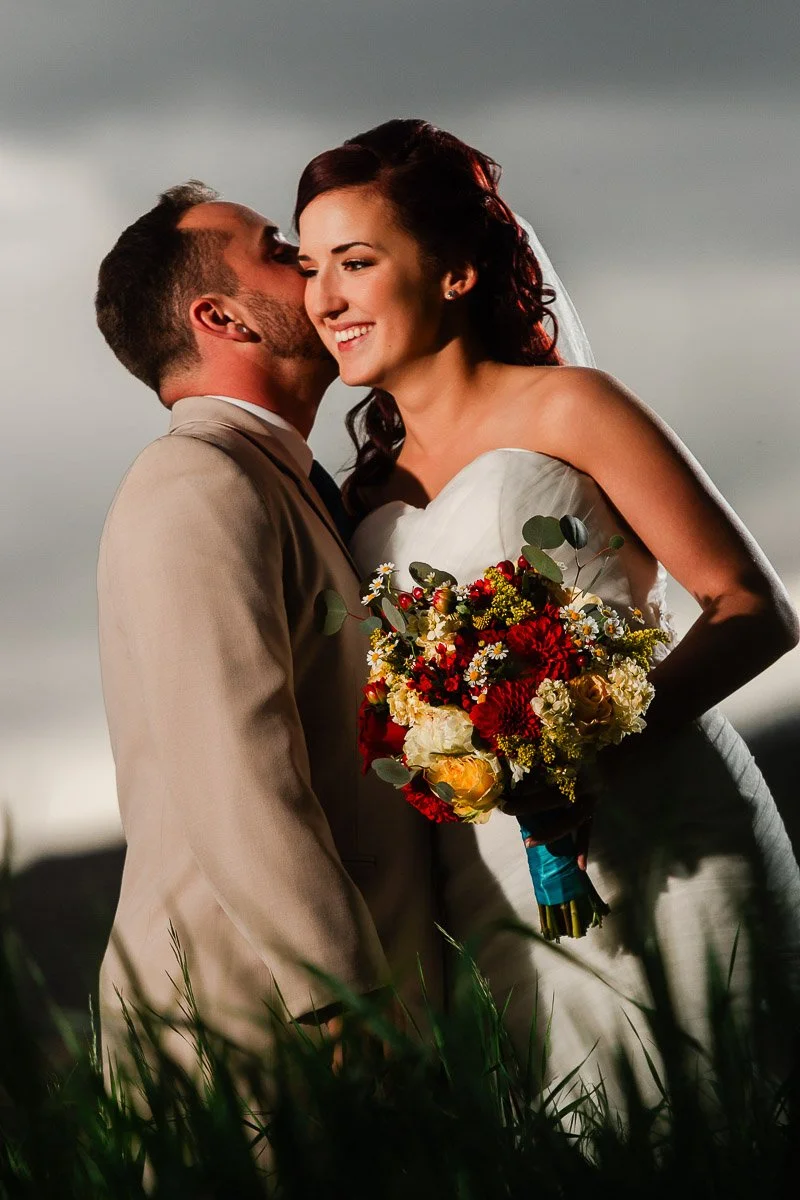 Bride in white dress holds a colorful bouquet while a groom in a beige suit whispers in her ear. They smile warmly against a cloudy sky backdrop.