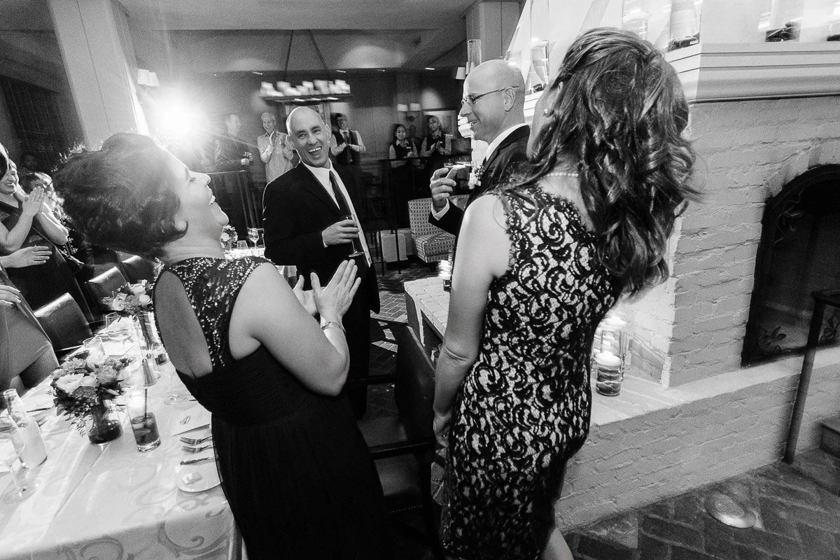 A lively party scene with elegantly dressed guests laughing and toasting near a table with flowers. The atmosphere is joyful and celebratory.