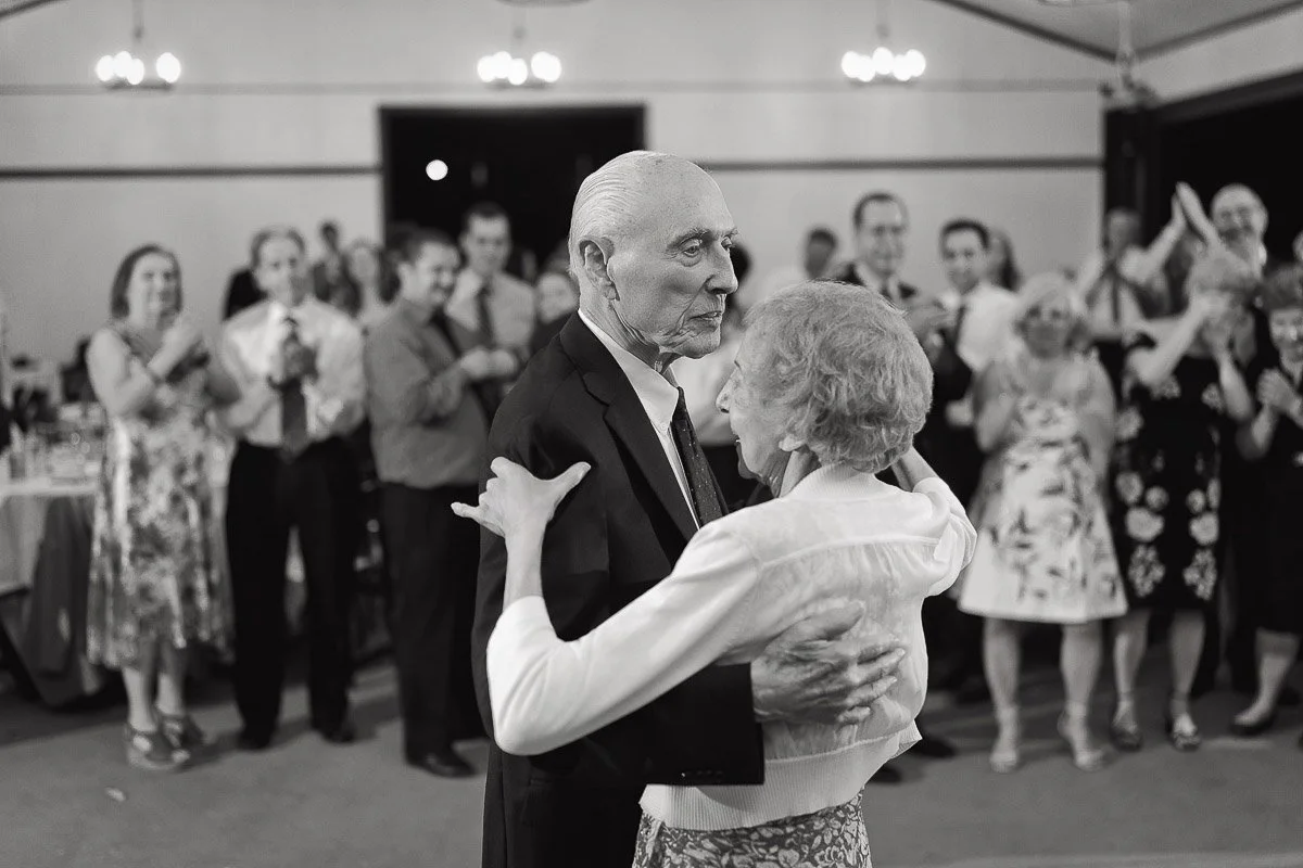 Elderly couple dancing together at an event, surrounded by smiling, clapping guests. The atmosphere is joyful and celebratory. Black and white.