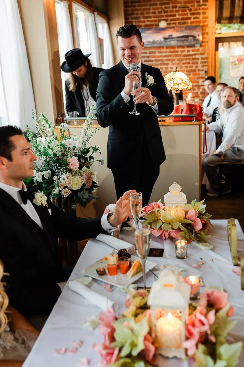 A man in a suit gives a toast at a wedding, smiling warmly. The table is decorated with flowers and lanterns, creating a festive and joyful atmosphere.