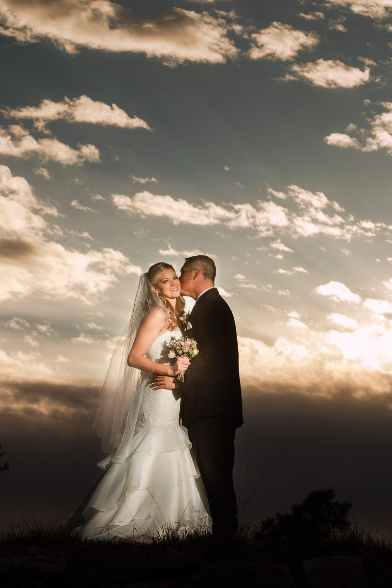 A bride and groom embrace at sunset, surrounded by a dramatic sky with scattered clouds. She smiles, holding flowers; his kiss conveys love and warmth during a Sanctuary Golf Course wedding in Sedalia, Colorado.