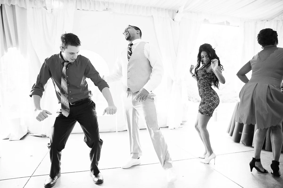 Guests joyfully dance at a lively indoor event. Two men in formal attire and two women in dresses express happiness and enjoyment on the dance floor.