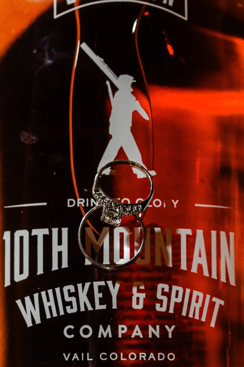 A diamond ring hangs on a bottle of 10th Mountain Whiskey & Spirit, highlighting its logo. Dark amber tones evoke warmth and elegance.