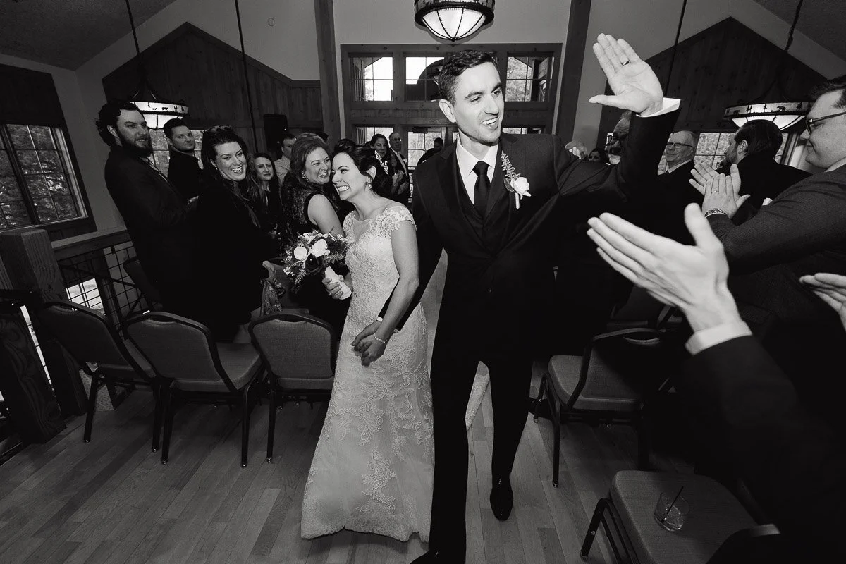 A joyful bride and groom walk down an aisle, holding hands. The groom high-fives a guest, while the bride smiles warmly. Guests clap and celebrate.