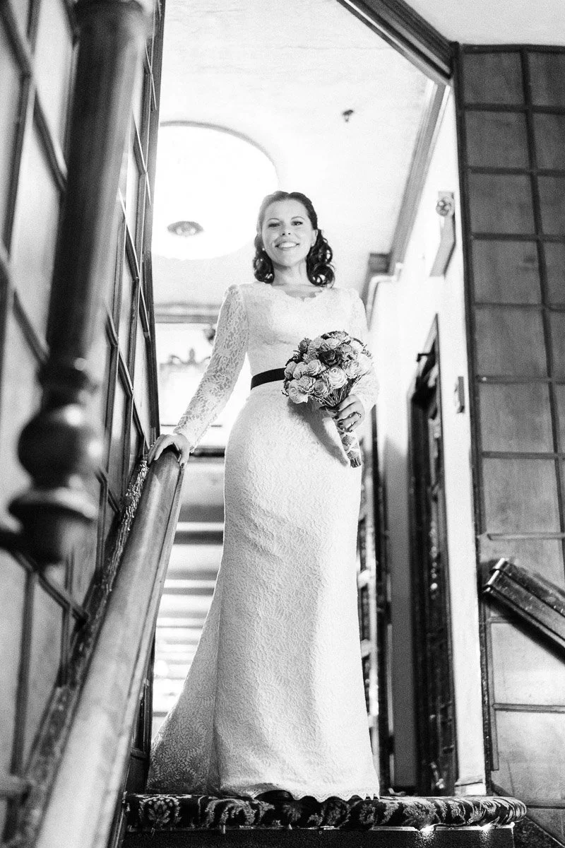 A woman in a lace wedding dress holds a bouquet, smiling while descending a staircase. The setting is elegant, with soft lighting and vintage decor.