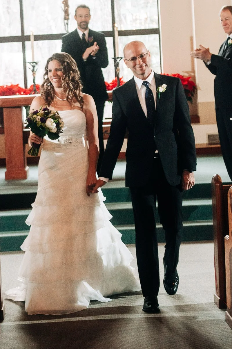 A newlywed couple walks hand-in-hand down a church aisle. The bride wears a tiered white gown and holds a bouquet. The groom is in a black suit. Guests clap joyfully.