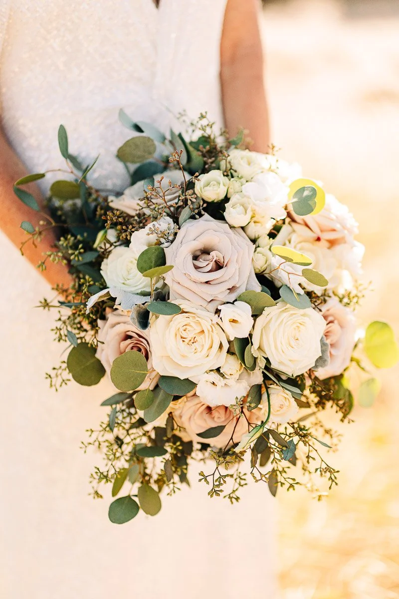 A person in a cream dress holds a bridal bouquet with white, blush roses, and greenery, conveying a romantic, elegant tone in warm sunlight.
