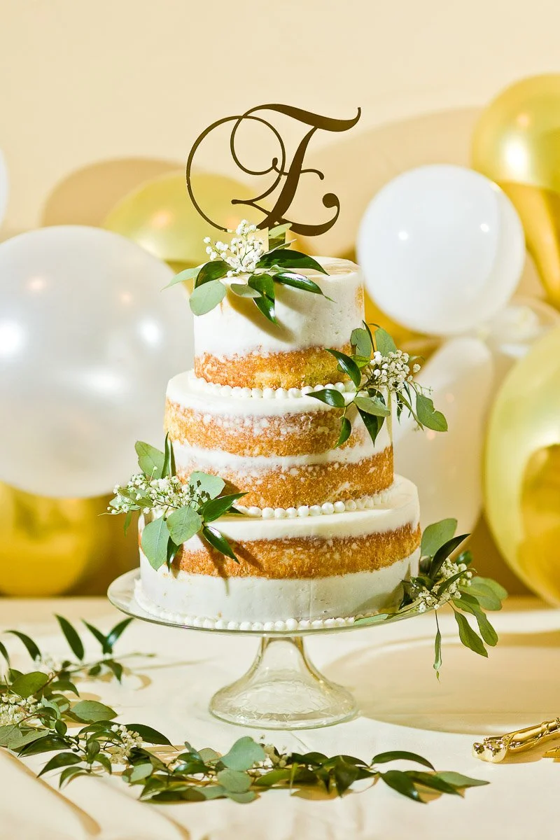 Three-tiered semi-naked cake with pearly white frosting, green leaves, and baby's breath. Topped with an ornate "F" against a backdrop of white and gold balloons. Elegant and festive.