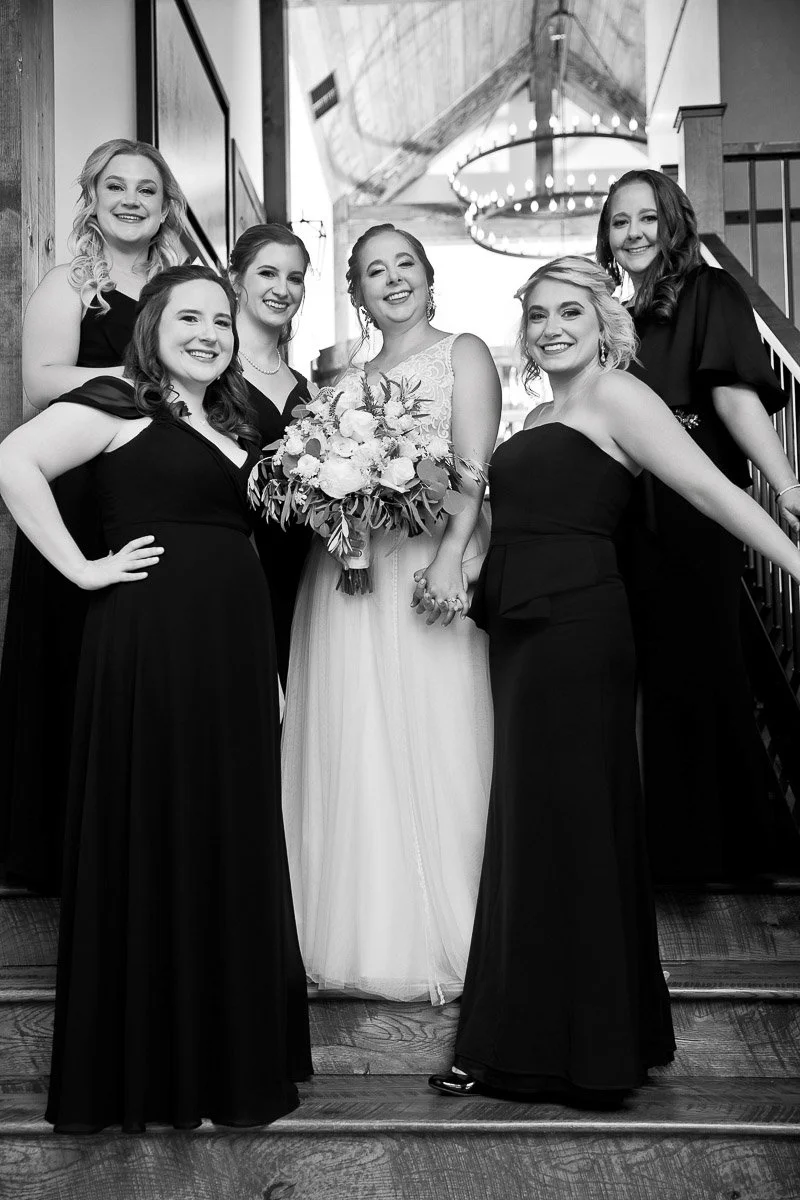 Black-and-white photo of a smiling bride with five bridesmaids. They pose on stairs, wearing elegant dresses, radiating joy and friendship.