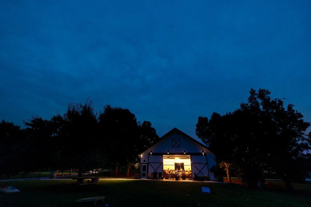 A barn with glowing interior lights stands amid dark trees under a deep blue evening sky. The scene conveys warmth and tranquility.