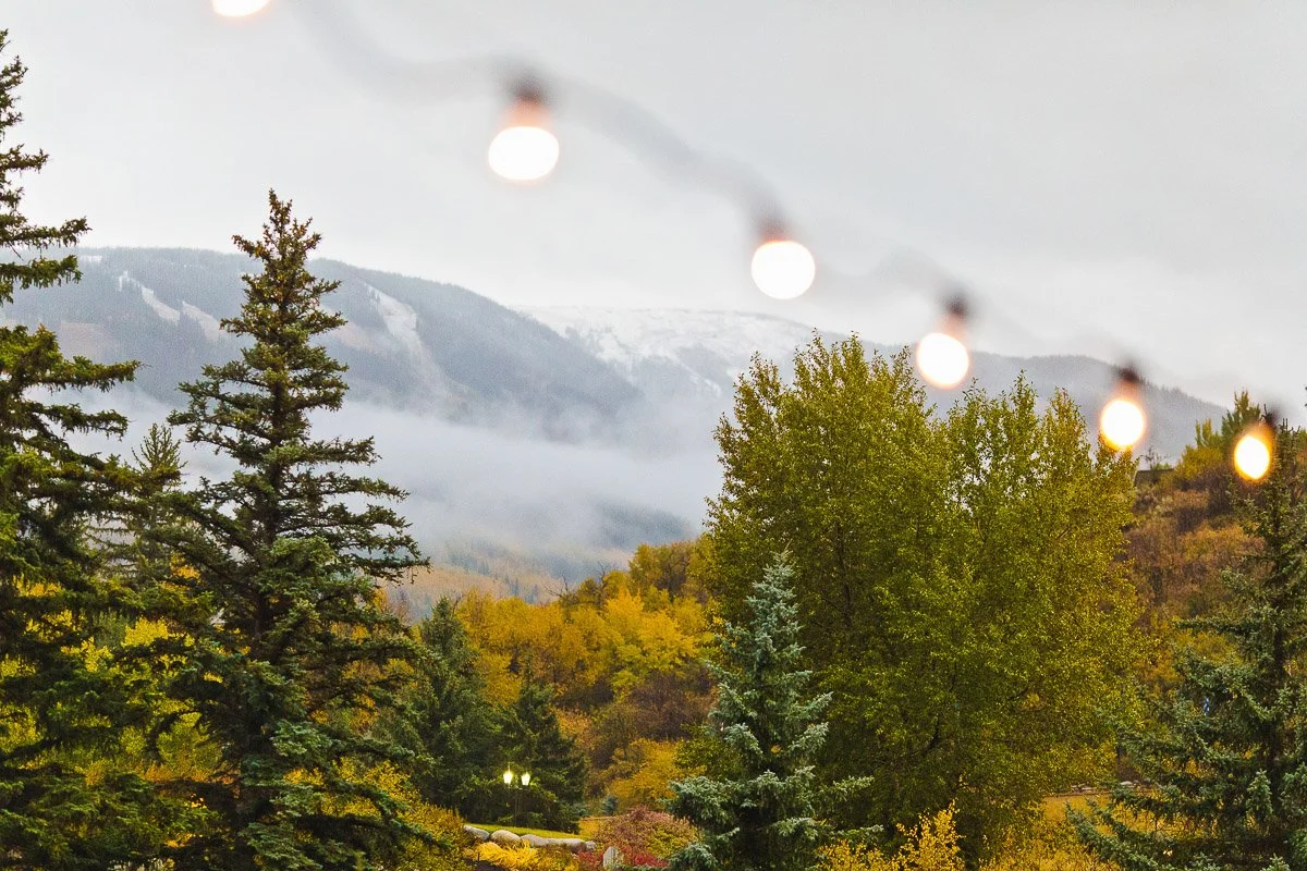 String lights hang over a misty mountain landscape with autumn trees. The scene is tranquil and cozy, blending nature's beauty with warm illumination.