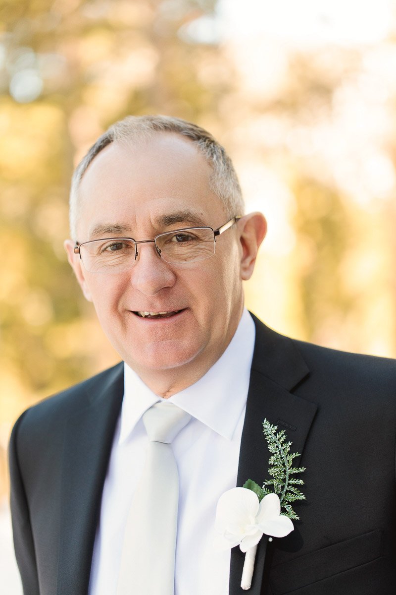 Elderly man in a suit with a white tie and boutonniere smiles warmly, standing outdoors with a soft, blurred background of greenery and sunlight.