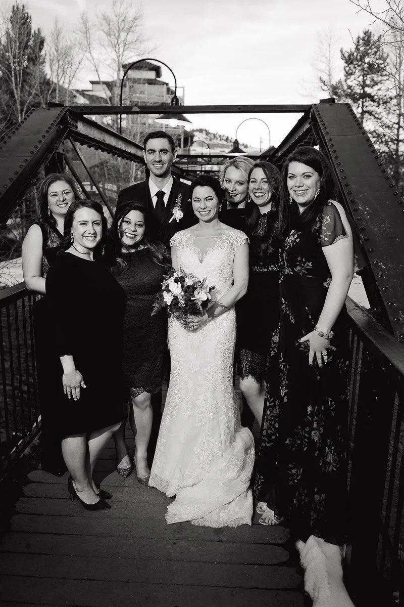 A joyful wedding party poses on a bridge. The bride in a lace dress holds flowers, surrounded by smiling friends dressed in formal attire. A serene, elegant scene.
