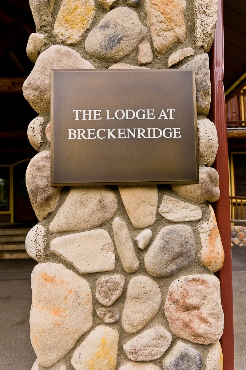 Stone pillar features a metal sign reading "The Lodge at Breckenridge" in white text, conveying a rustic and welcoming ambiance.