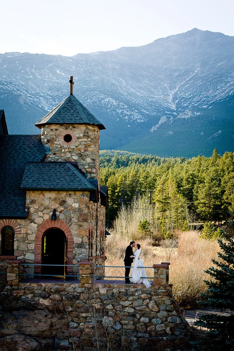 A couple in wedding attire stands on a stone balcony of a rustic chapel, nestled among dense pine forests with majestic mountains in the background. Romantic and serene.